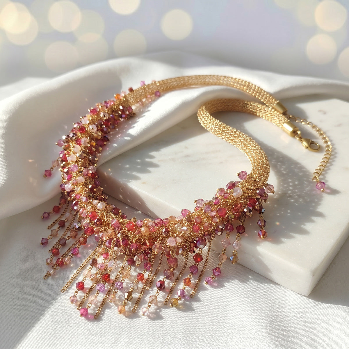 A gold-tone statement necklace with cascading pink, red, and peach faceted beads, displayed on a white marble block over a white fabric background.