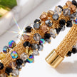 A close-up of a gold-toned braided bracelet adorned with faceted crystal beads in black, clear, brown, and iridescent colors, shown on a light neutral background with a bright lens flare effect.