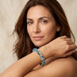 A woman with long brown hair wearing a beaded bracelet with blue, turquoise, and gold beads, posing against a neutral beige background.