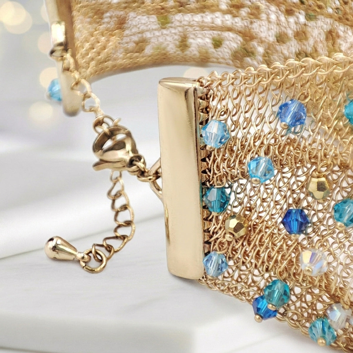 Close-up of a gold-tone mesh clutch bag with blue faceted rhinestones and a gold clasp, resting on a white surface.