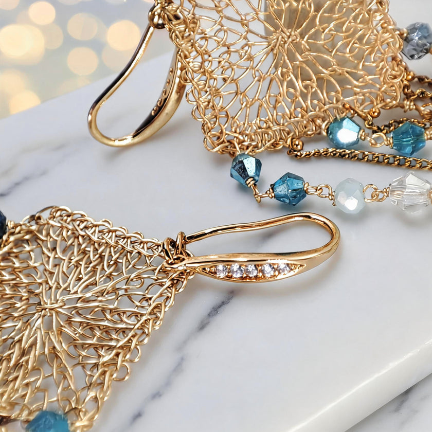 Gold drop earrings with a diamond-shaped handknitted wire mesh square panel and cascading strands of blue reflective crystals.

