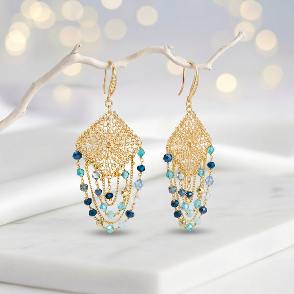 Gold drop earrings with a diamond-shaped handknitted wire mesh square panel and cascading strands of blue reflective crystals.
