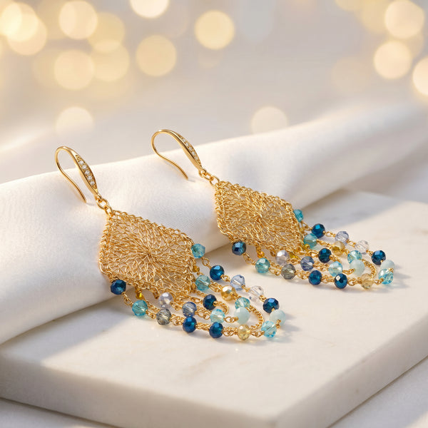 Gold drop earrings with a diamond-shaped handknitted wire mesh square panel and cascading strands of blue reflective crystals.
