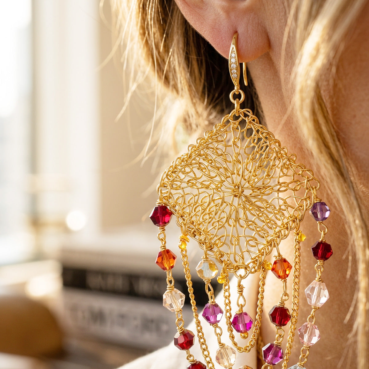 A close-up of a woman's ear wearing a large gold dangle earring with intricate filigree detailing and dangling faceted beads in red, orange, purple, and clear crystal.