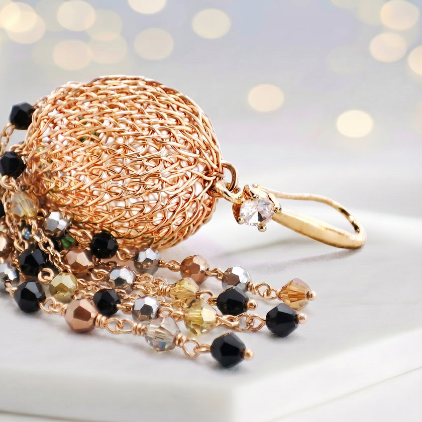 Gold drop earrings featuring a sphere-shaped handknitted wire shape and tassel strands of black and bronze crystals.