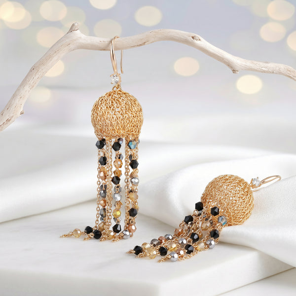 Gold drop earrings featuring a sphere-shaped handknitted wire shape and tassel strands of black and bronze crystals.