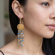 A close-up of a woman wearing a dangling tassel earring. The earring features a gold-toned hook, a round woven gold top, and multiple strands of blue, light blue, and clear faceted beads.