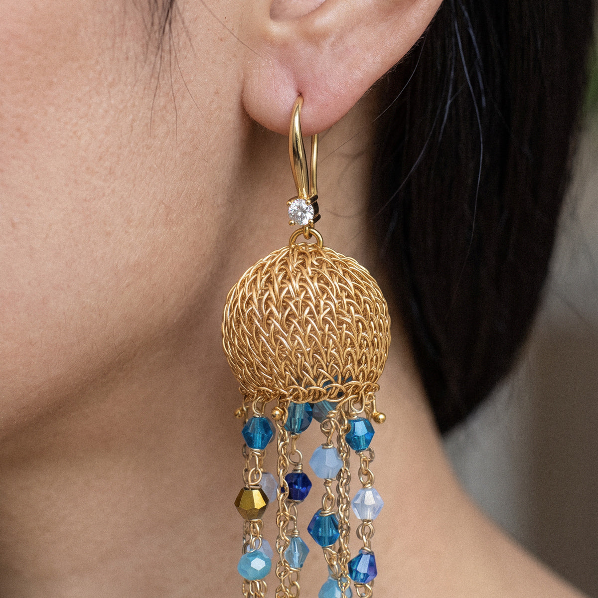 A close-up of a gold dangle earring worn on a person's ear, featuring a textured gold mesh globe and dangling faceted blue beads.