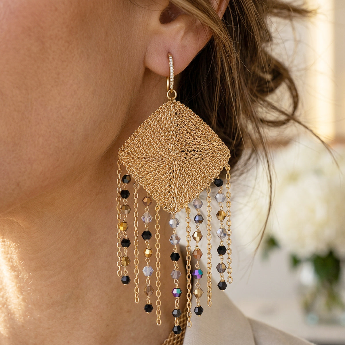 A close-up of a model wearing a large gold-toned dangle earring with a woven diamond-shaped pendant and hanging beaded chains.
