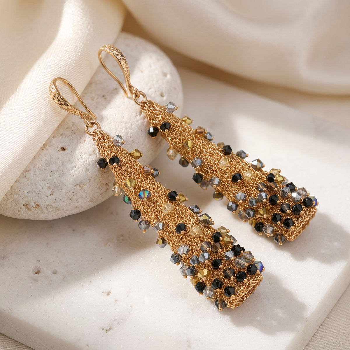 A pair of gold-toned drop earrings with textured mesh and dangling faceted crystal beads in black, gold, and iridescent tones, styled on a light stone slab with soft cream fabric.