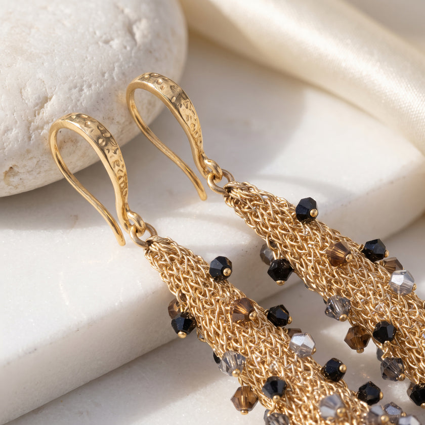 Gold-tone dangle earrings with textured fishhook backs, woven mesh strands, and small faceted black and smoky brown beads, displayed on a light stone and satin fabric background.