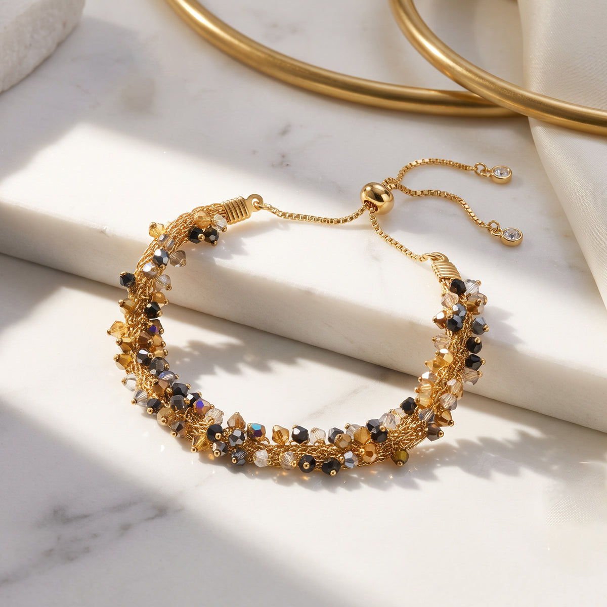 A gold-toned beaded bracelet with clear and black faceted beads, styled on a white marble surface with draped white fabric and gold hoop earrings in the background.