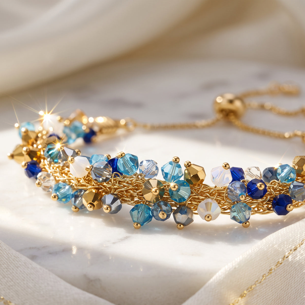 A gold chain bracelet adorned with faceted blue, light blue, and white crystals, displayed on a draped cream fabric surface.