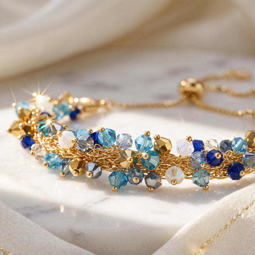 A gold chain bracelet adorned with faceted blue, light blue, and white crystals, displayed on a draped cream fabric surface.
