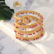 A stack of gold-toned beaded bracelets arranged on a white marble surface with a white orchid and green leaf in the background. The bracelets feature woven gold bands and rows of small faceted beads in red, pink, purple, and clear tones.