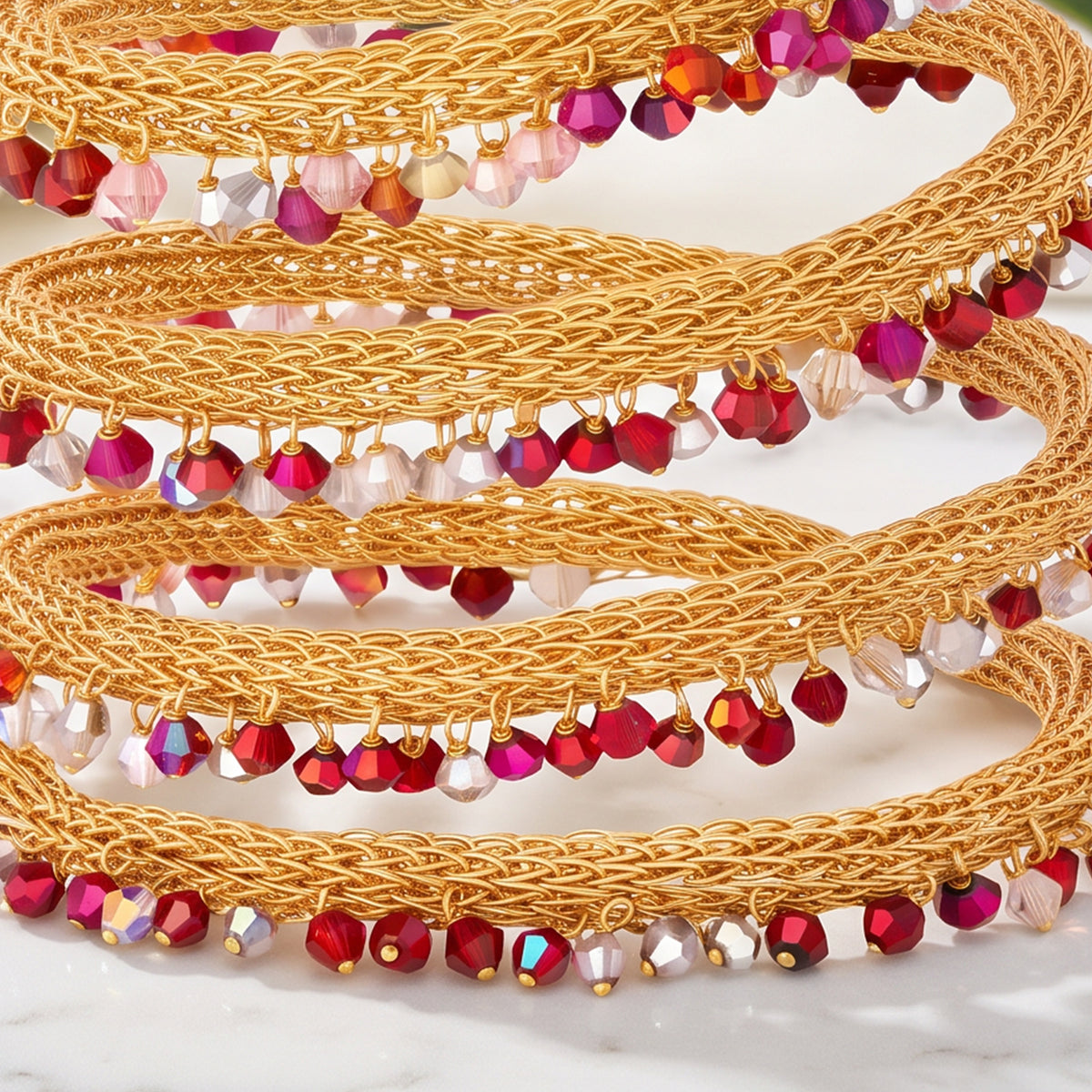 A close-up of a gold braided beaded belt or waist wrap, coiled on a light marble surface. The belt features woven gold cord and is decorated with dangling faceted beads in shades of red, magenta, pink, and clear iridescent.