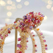 A close-up of a gold-toned beaded headband with a floral crown design, decorated with faceted pink, red, purple, and orange crystals, displayed on a stand against a soft, blurred bokeh background.