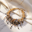 A gold-toned beaded statement necklace with cascading crystal drops in blue, black, and gold, styled on draped cream satin fabric over a white marble surface.
