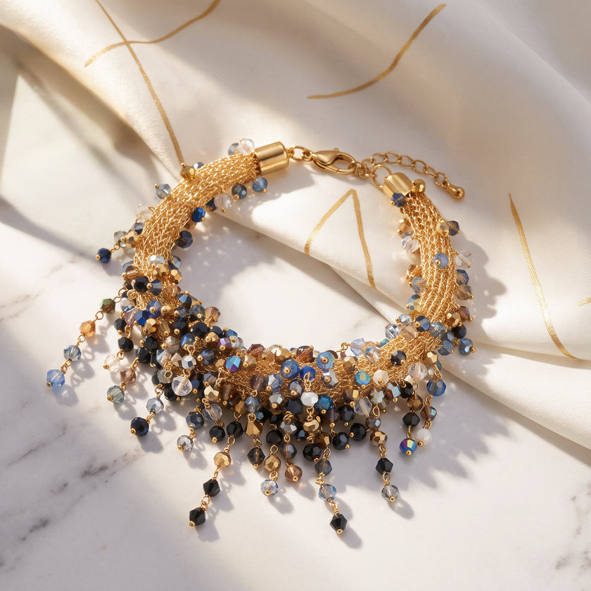 A gold-toned beaded statement necklace with cascading crystal drops in blue, black, and gold, styled on draped cream satin fabric over a white marble surface.