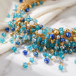 A close-up of a beaded necklace with cascading blue faceted beads, gold wire wrapping, and a few white beads, styled on a white fabric and marble surface.
