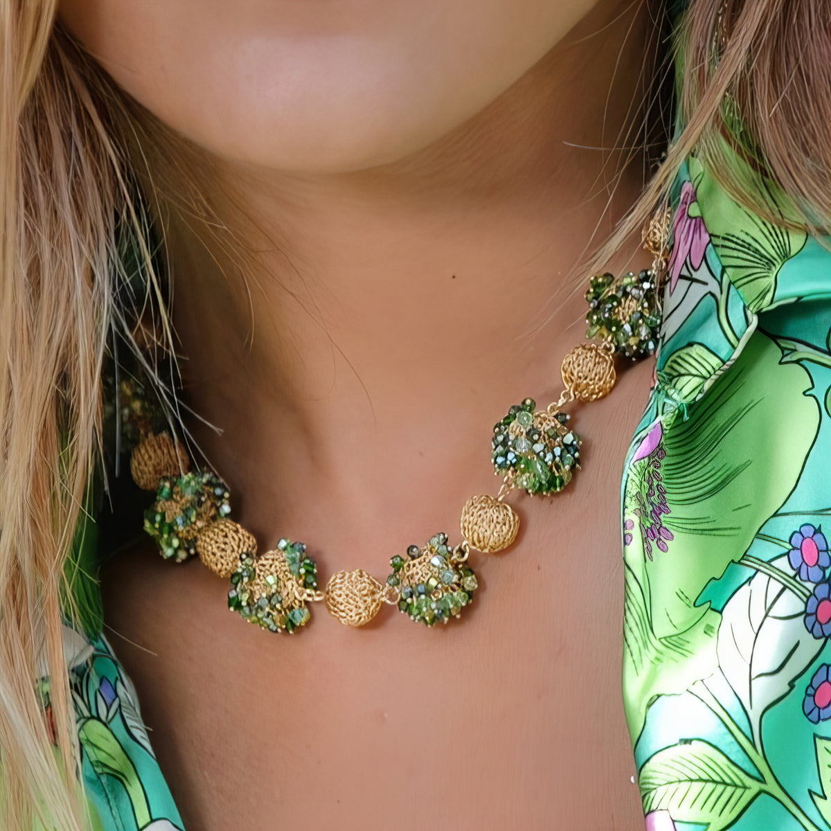 Close-up of a woman wearing a gold and green necklace with floral earrings against a green patterned background.