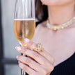 A close-up lifestyle shot of a woman holding a glass of champagne, showcasing a detailed gold ring with clear crystals on her finger. She is also wearing a sparkling gold crystal necklace.
