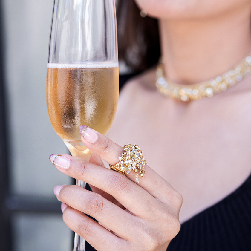 A close-up lifestyle shot of a woman holding a glass of champagne, showcasing a detailed gold ring with clear crystals on her finger. She is also wearing a sparkling gold crystal necklace.