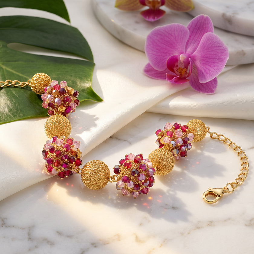 A gold chain bracelet with decorative floral beads, styled on a white marble surface with a green leaf and a pink orchid flower.