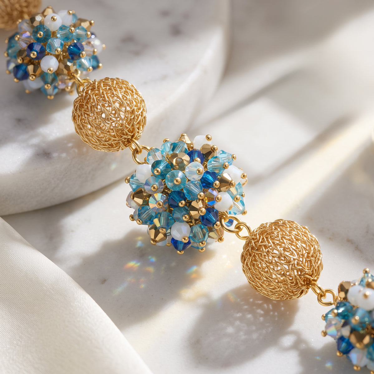 Gold beaded statement earrings with textured spherical gold beads and blue, light blue, and white faceted crystal clusters, displayed on a white marble slab with soft white fabric.