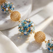 Gold beaded statement earrings with textured spherical gold beads and blue, light blue, and white faceted crystal clusters, displayed on a white marble slab with soft white fabric.