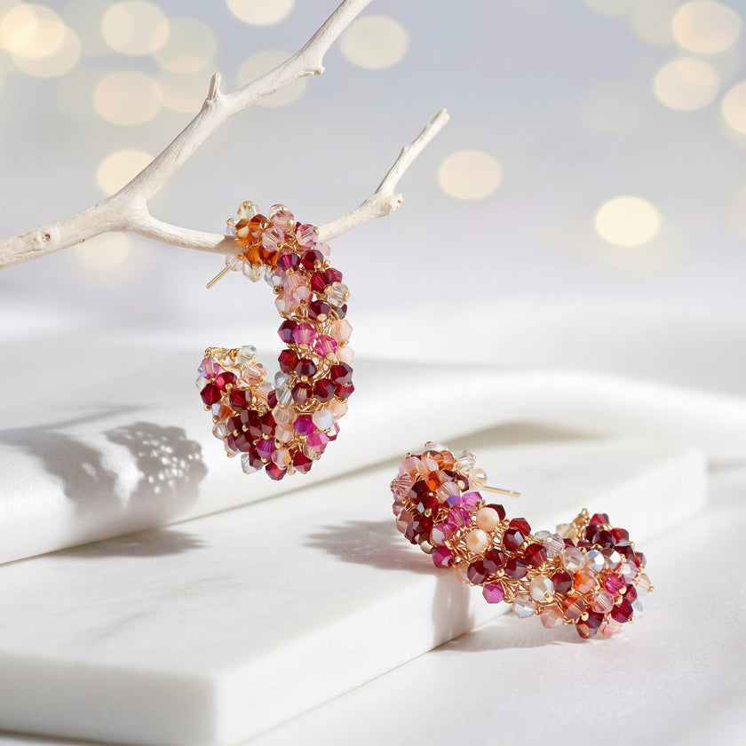 Gold loop earrings displaying a sunburst handknitted wire mesh frame filled with hanging pink and red crystals.
