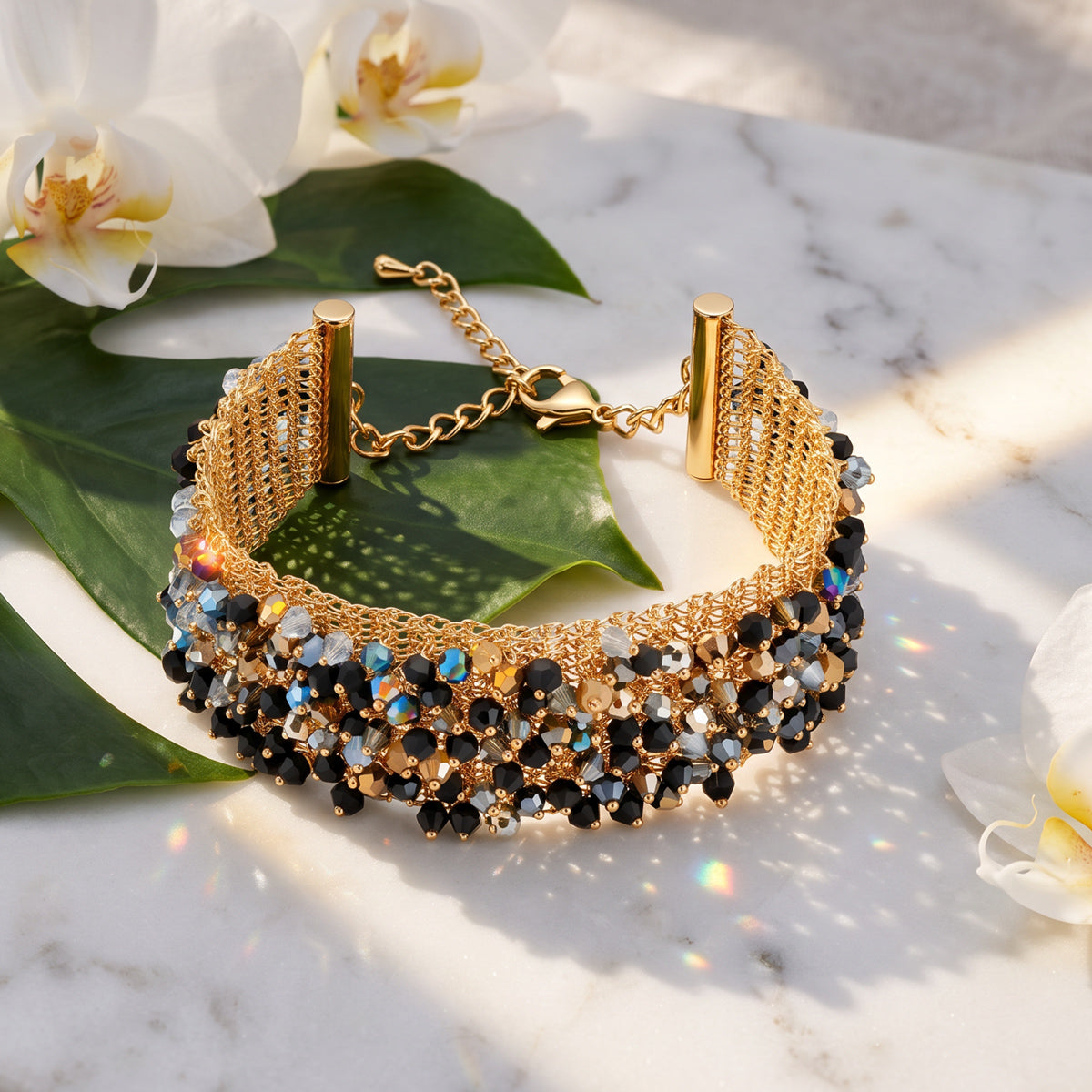 A wide gold-tone beaded cuff bracelet with black, white, and iridescent faceted beads, displayed on a white marble surface with green leaves and white orchids.
