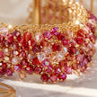 A close-up of a gold-tone beaded bracelet or cuff covered with faceted crystal beads in shades of pink, magenta, and red, resting on a light marble-like surface.