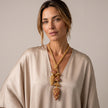 A woman wearing a beige satin blouse and a chunky gold-toned statement necklace with starfish and shell-like beads, photographed against a plain warm beige studio background.